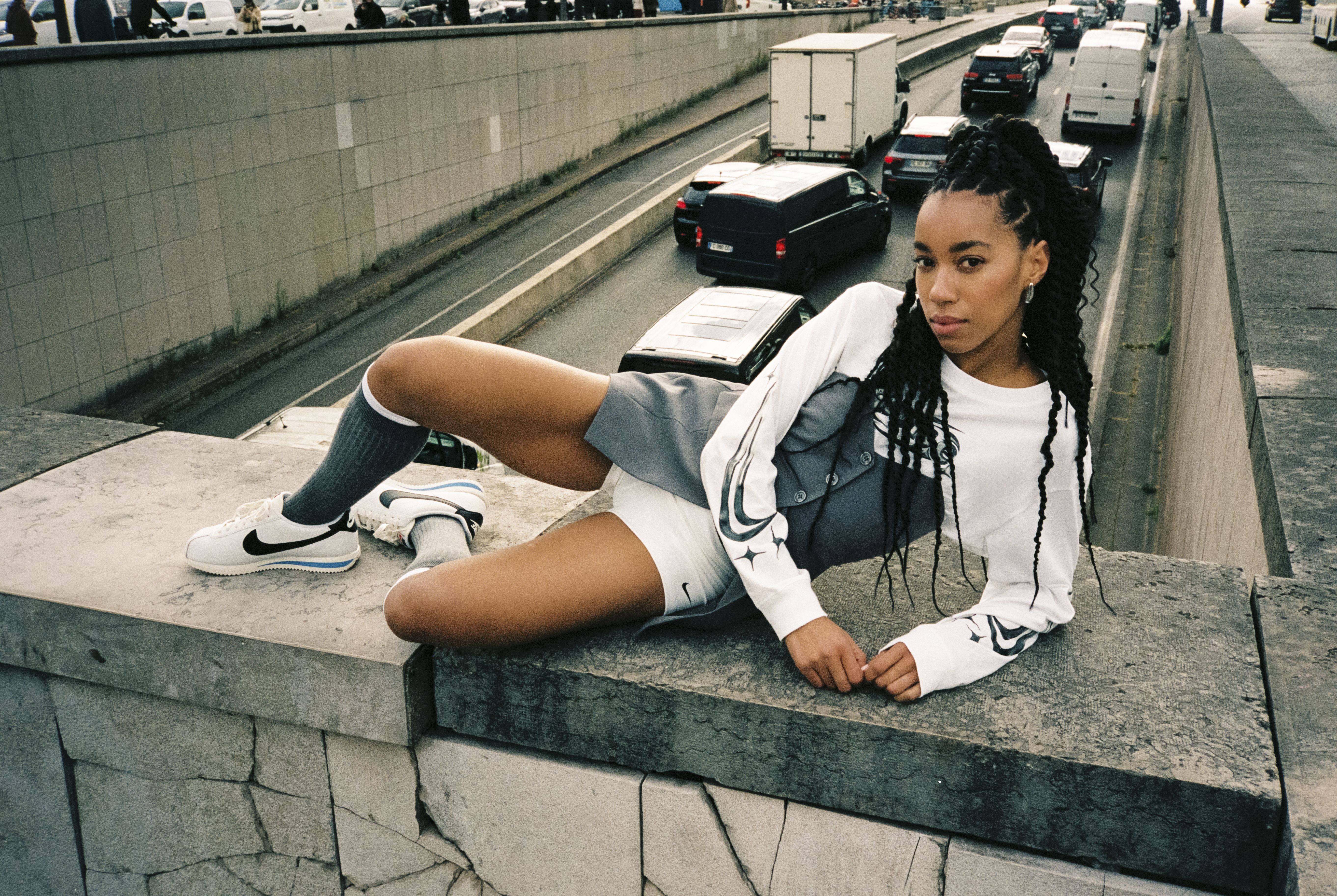 Γυναίκα με λευκό πουλόβερ NIKE και λευκά παπούτσια NIKE ποζάρει σε τσιμεντένιο τοίχο πάνω από πολυσύχναστο δρόμο